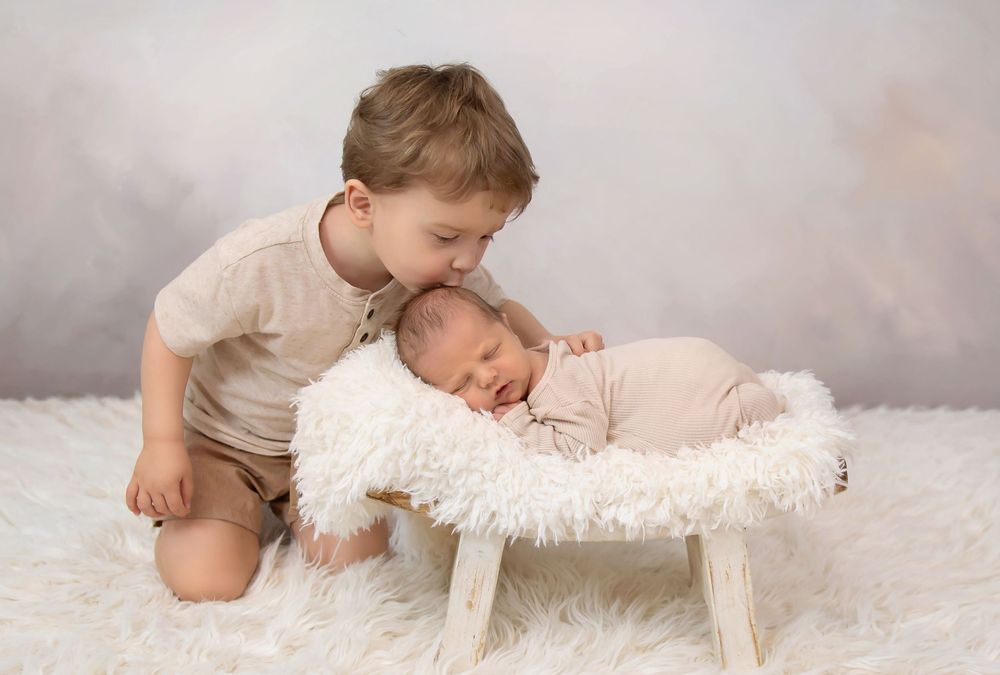 Older toddler boy in beige outfit kissing sleeping newborn baby in white fluffy basket during professional studio photo session.