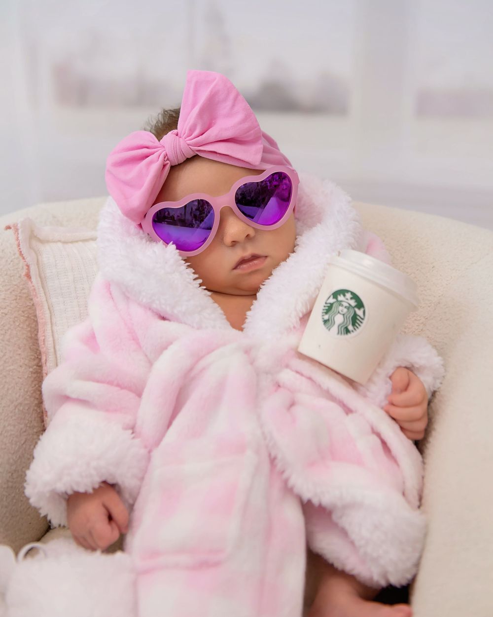 Adorable newborn baby in pink plaid robe and heart sunglasses holding a Starbucks cup with pink bow headband.