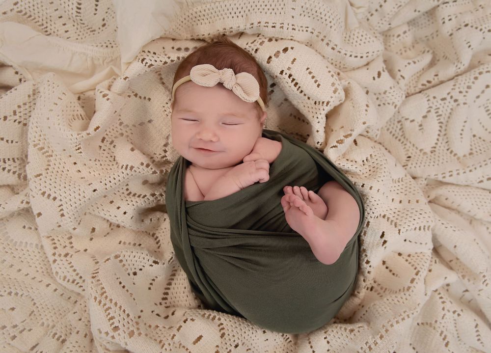 Newborn baby wrapped in olive green swaddle with bow headband, sleeping peacefully on cream lace blanket.