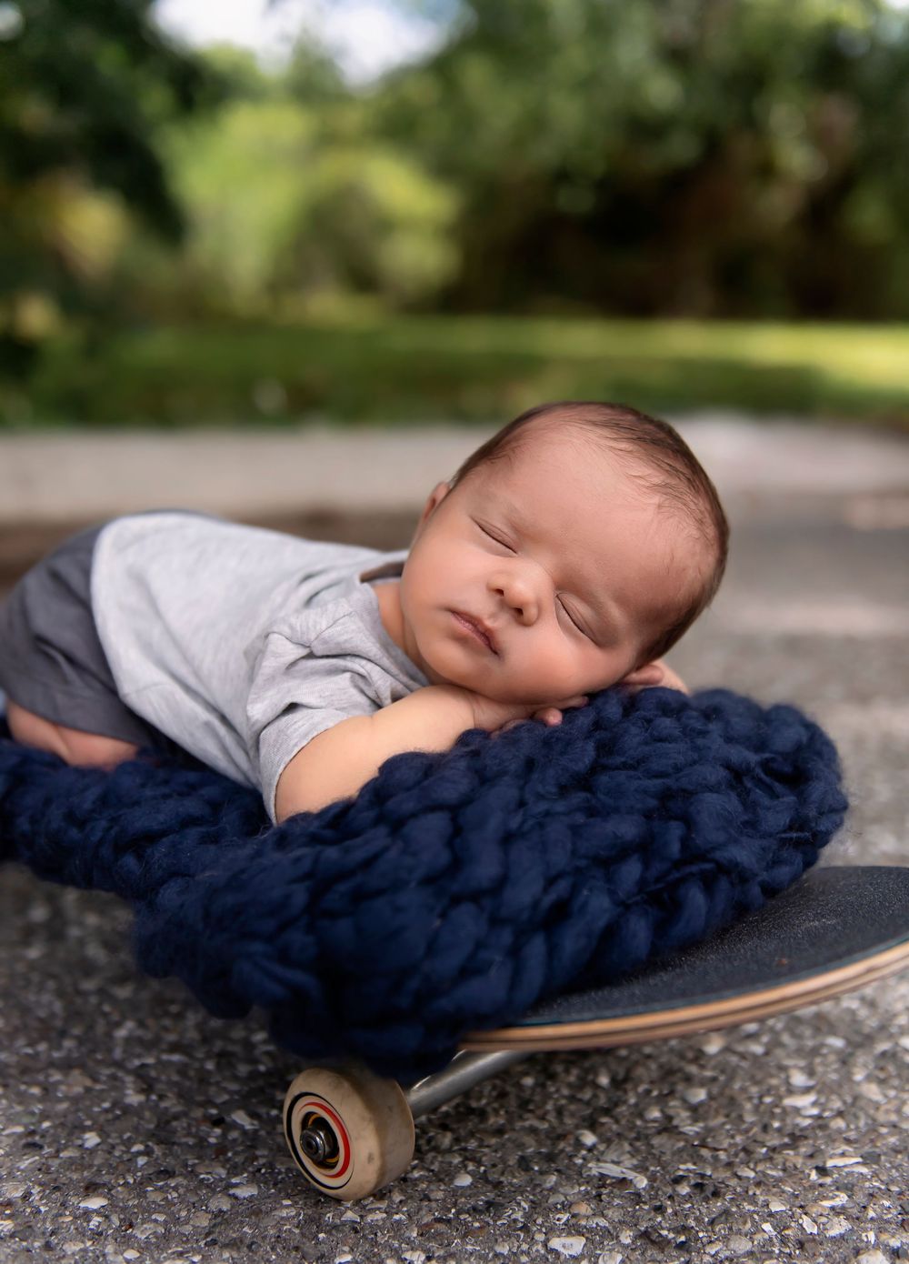 Sleeping newborn baby resting on a navy blue chunky knit blanket on a skateboard outdoors.