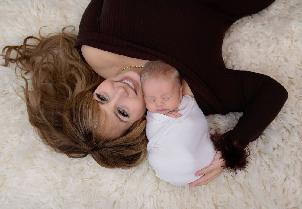 A smiling mother in a brown dress lies on a fluffy white rug, cradling her swaddled newborn baby.