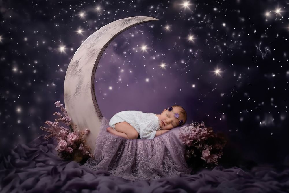 Newborn baby sleeping peacefully on a crescent moon prop surrounded by stars and pink flowers in a dreamy purple setting.