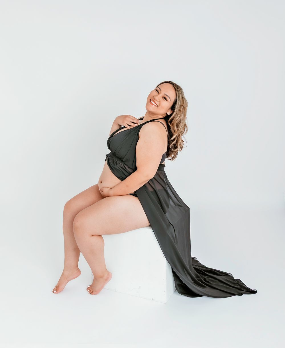 Pregnant woman wearing a sheer black gown sitting on a white cube with white background at studio of SC Photography Studio in Angleton TX