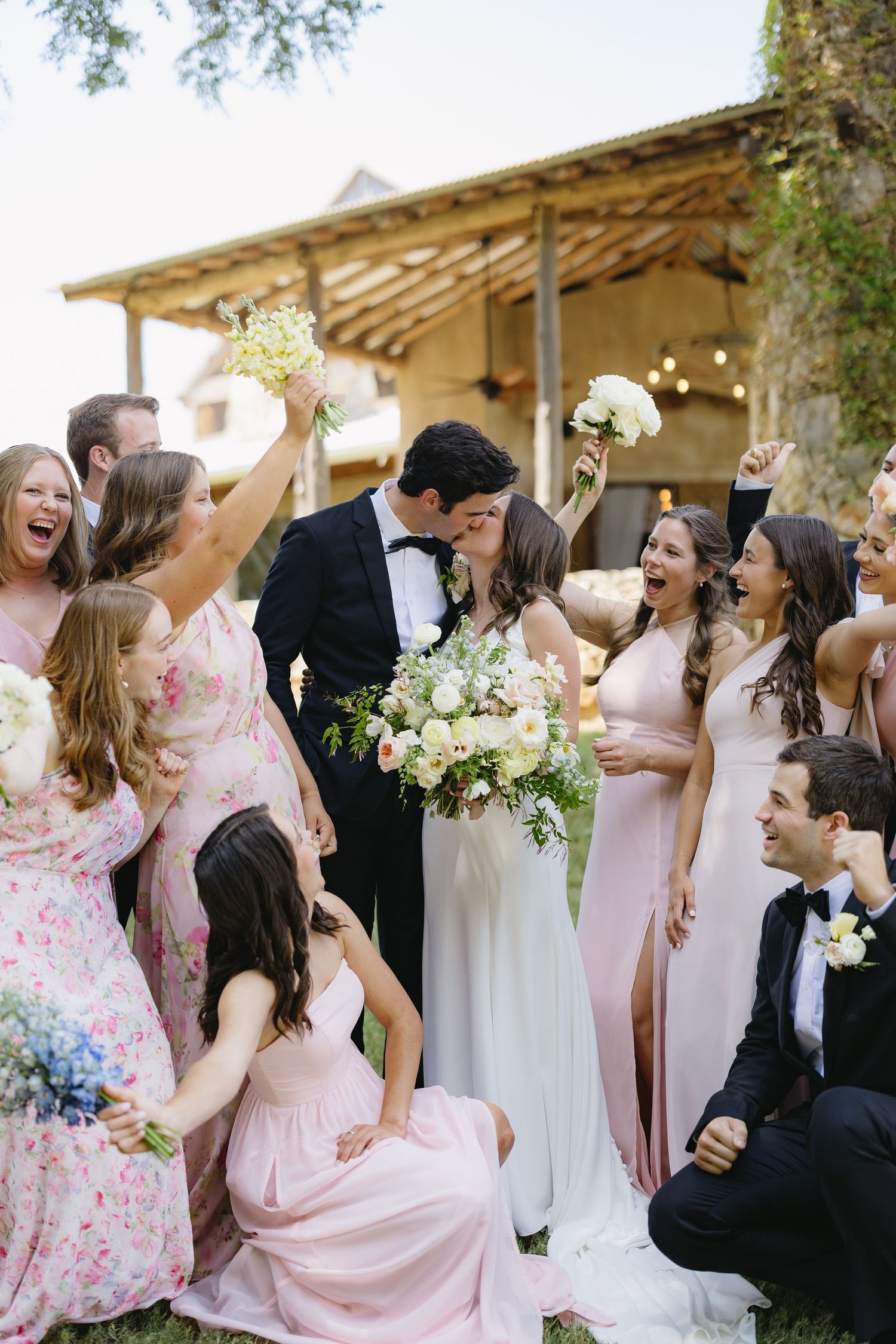Joyful wedding party celebrating with bride and groom kissing, bridesmaids in pink dresses holding bouquets.