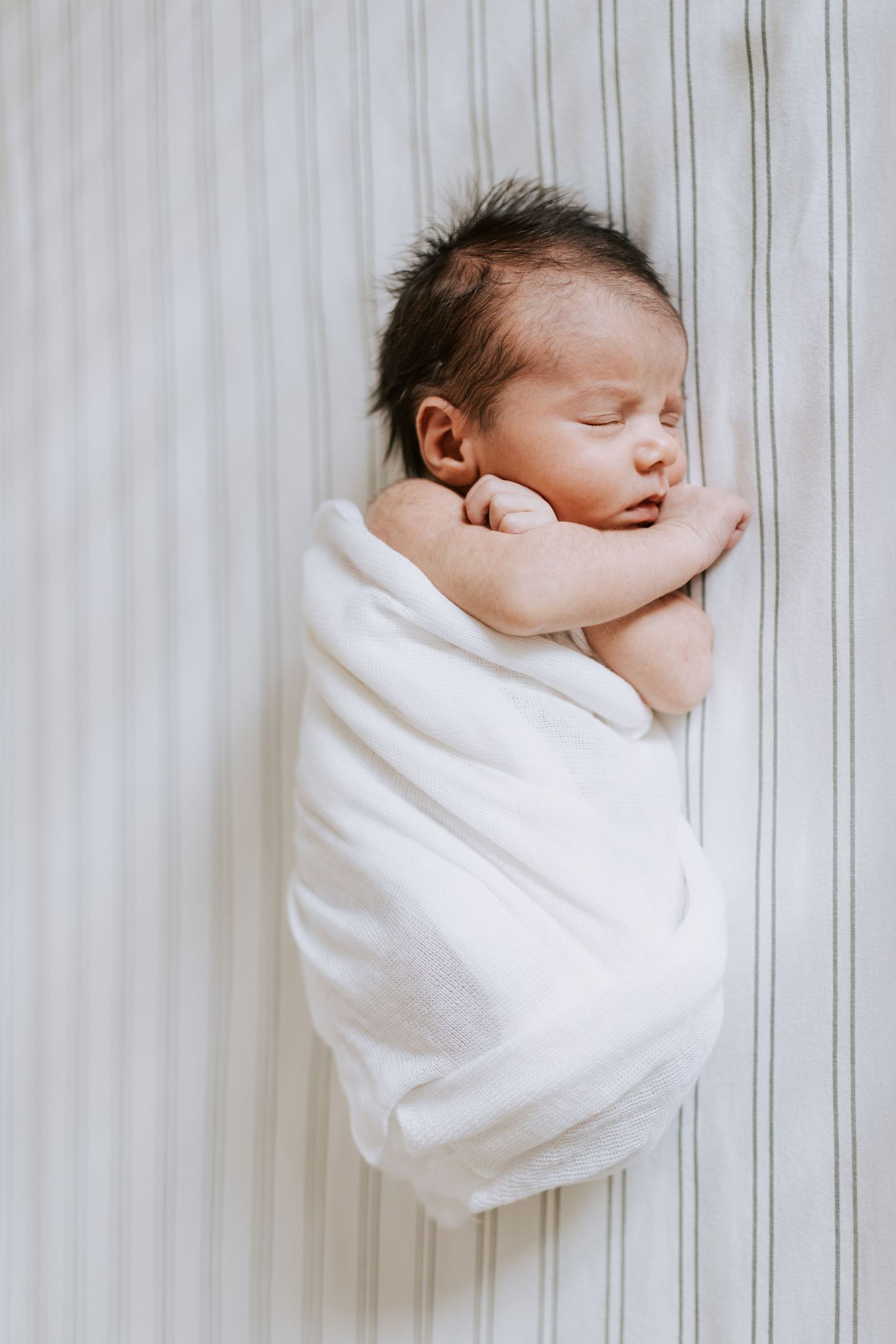Sleeping newborn baby wrapped in soft white blanket against striped background.