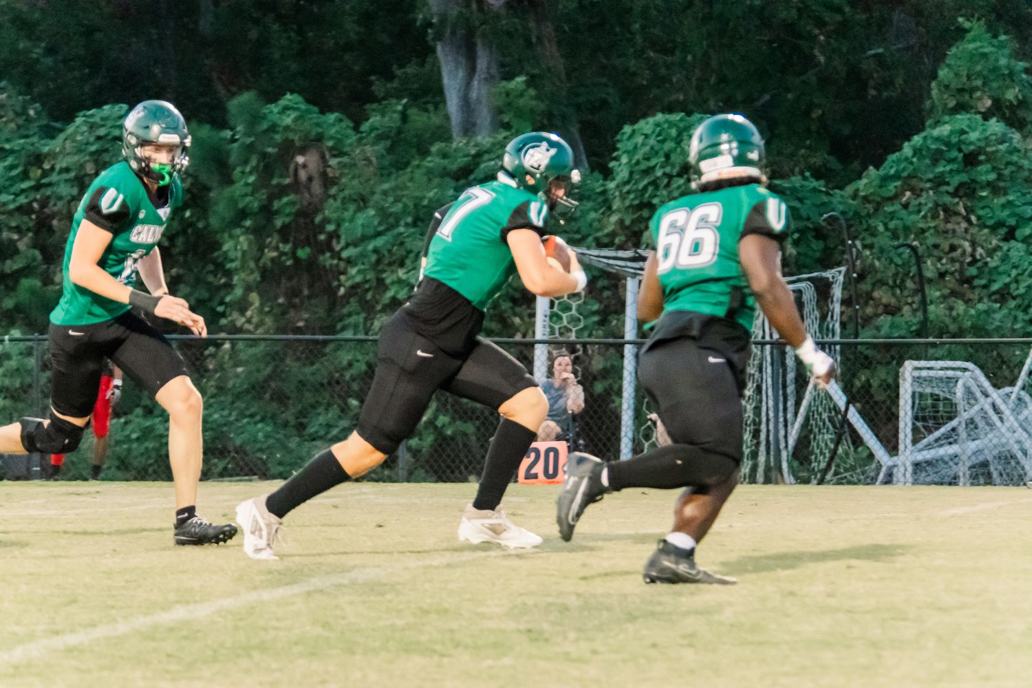 Three football players in green uniforms practice plays on a field during an evening game or practice session.