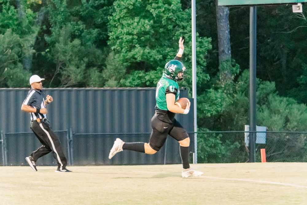 Football player in green uniform celebrates with raised arm on field, referee and trees visible in background.