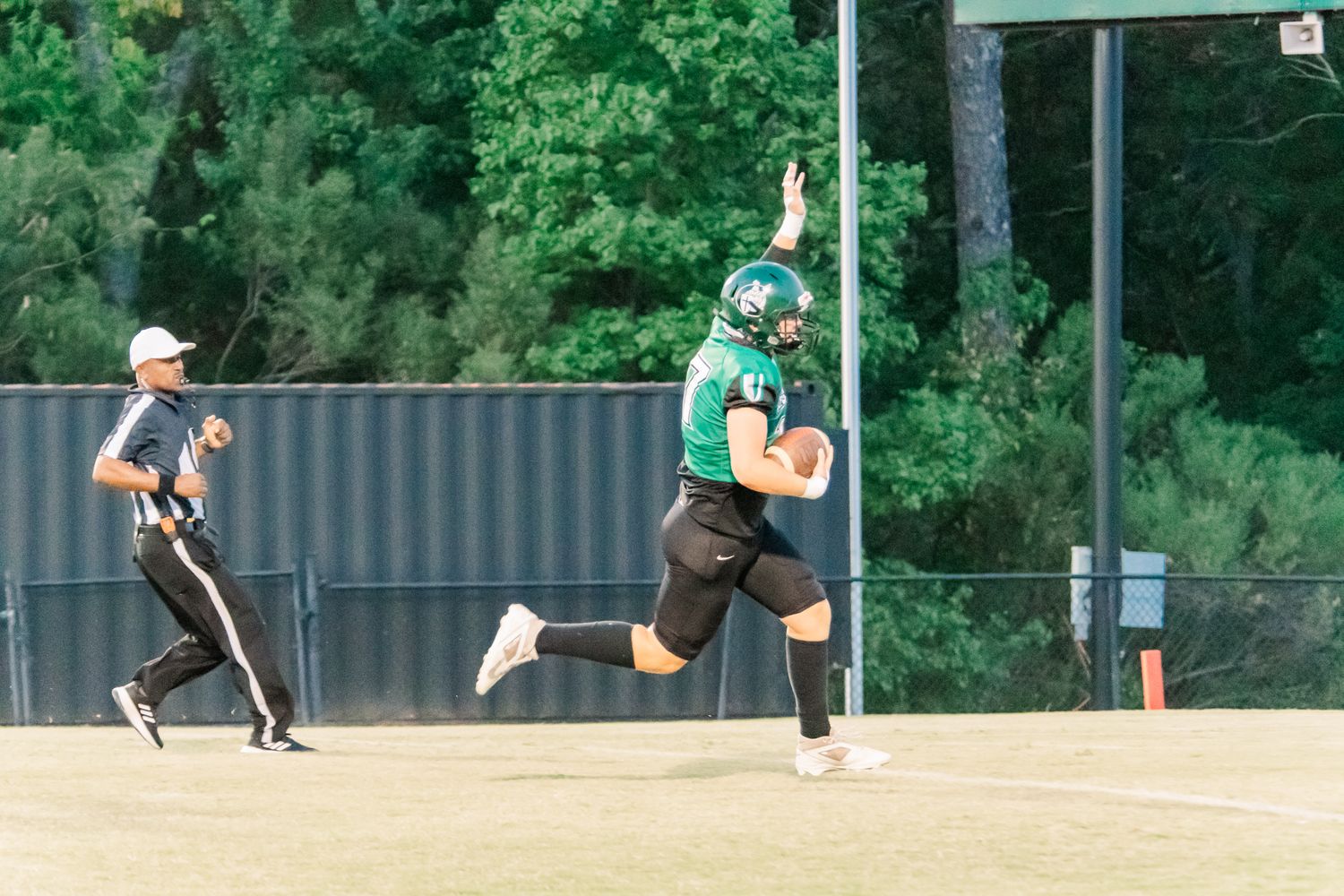 Football player in green uniform celebrates with raised arm on field, referee and trees visible in background.