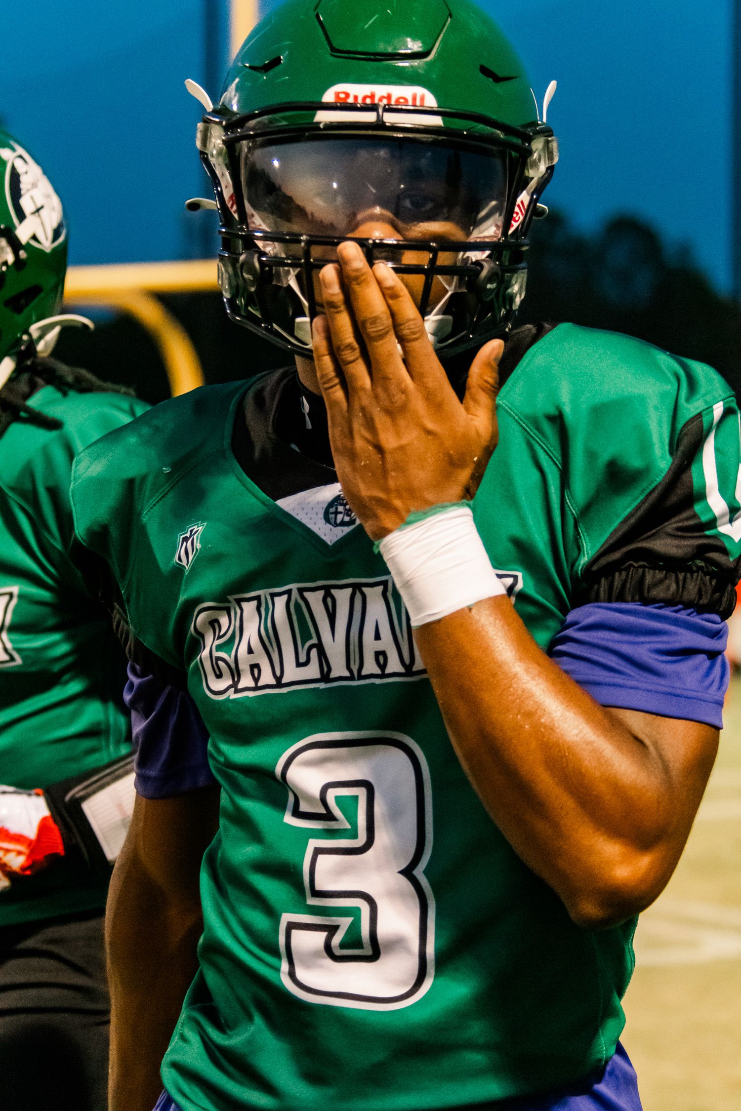 Calvary football player #3 in green jersey and Riddell helmet covers mouth on sideline during game.