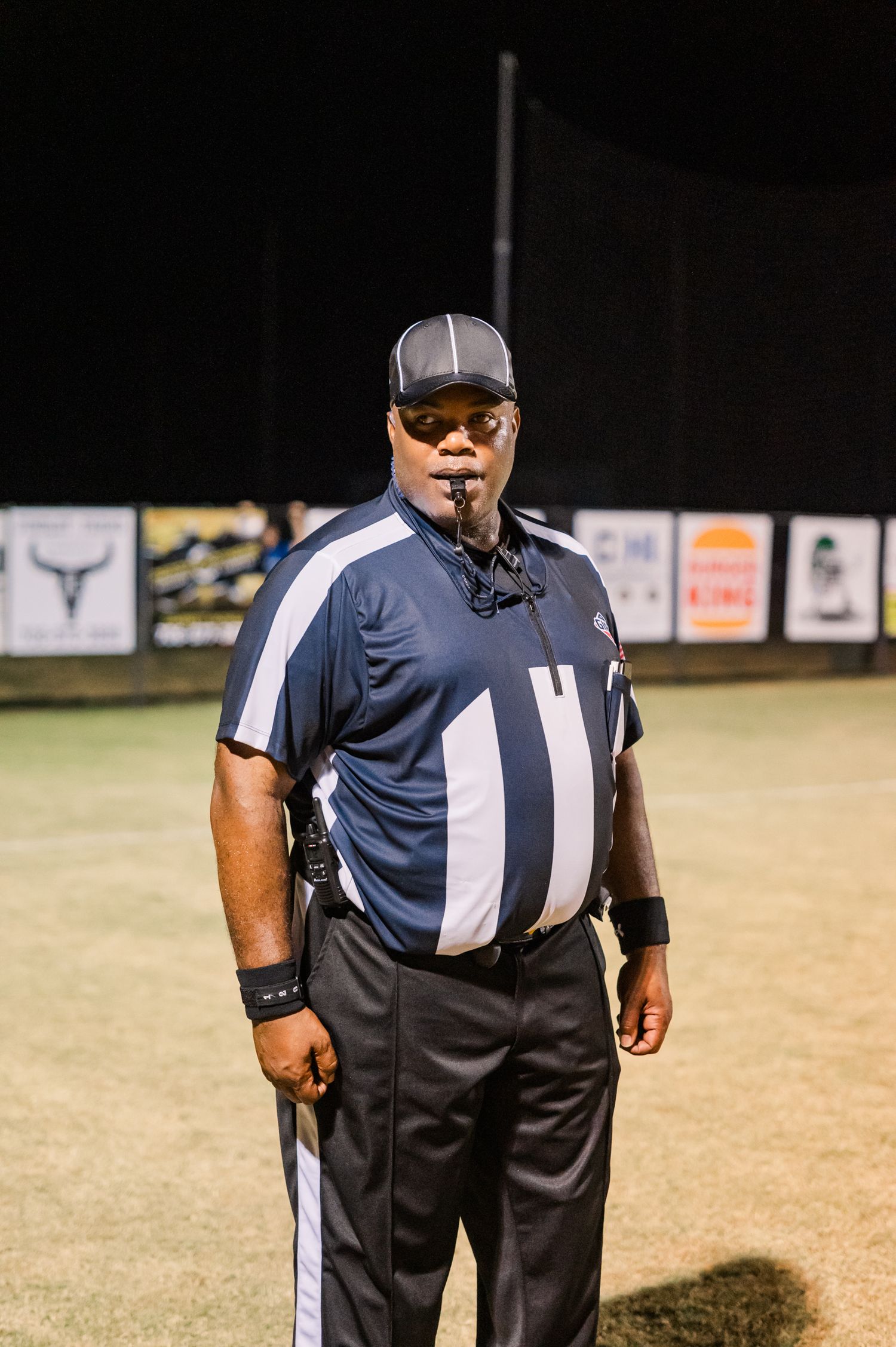 A football referee in navy blue and white striped uniform stands on the field at night with a whistle in his mouth.
