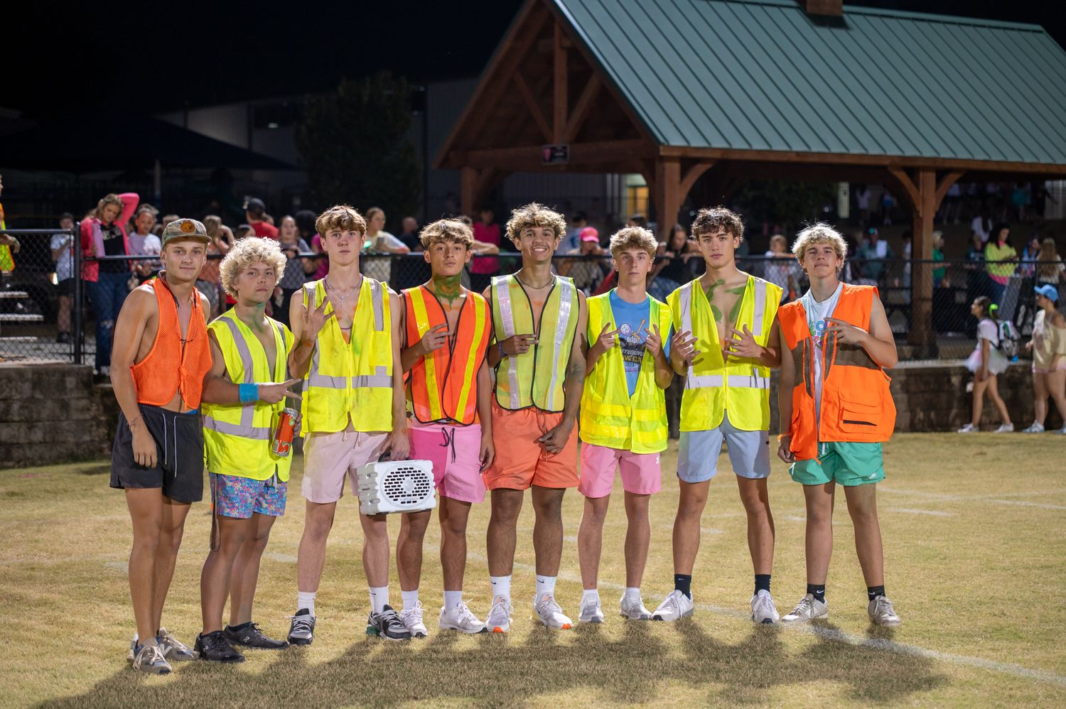 Group of teenage boys wearing neon yellow safety vests and colorful shorts posing together outdoors at a summer event.