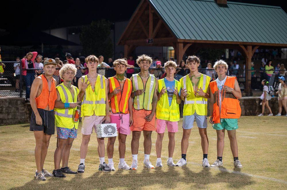 Group of teenage boys wearing neon yellow safety vests and colorful shorts posing together outdoors at a summer event.