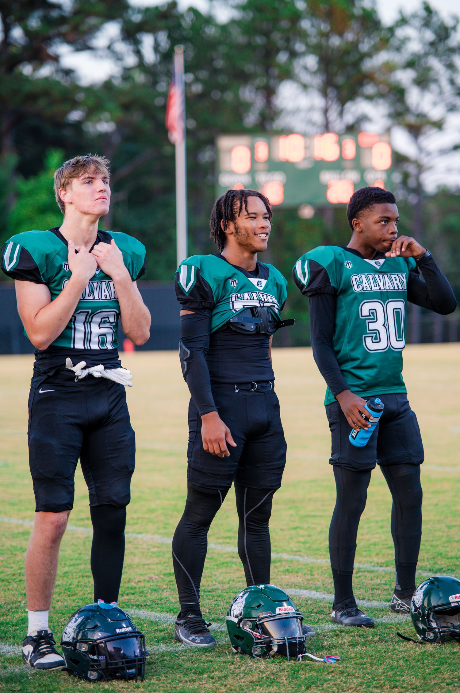 Three Calvary football players in green uniforms stand on the sideline during a game, wearing numbers 16 and 30.