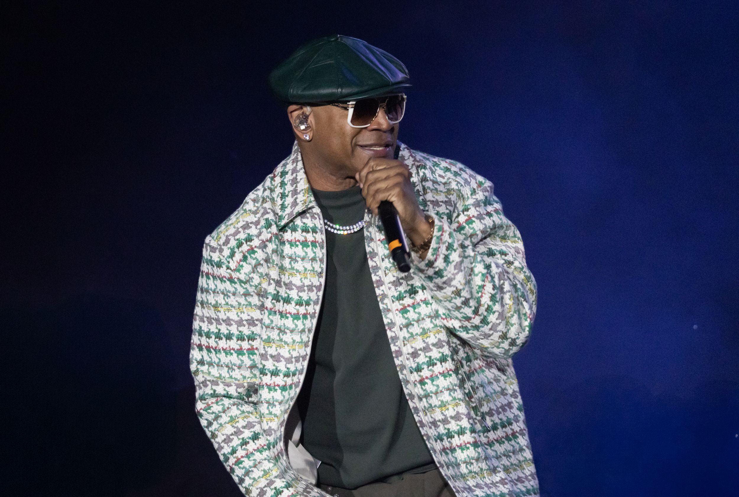 A performer wearing a patterned white and black jacket and dark cap stands against a deep blue background.