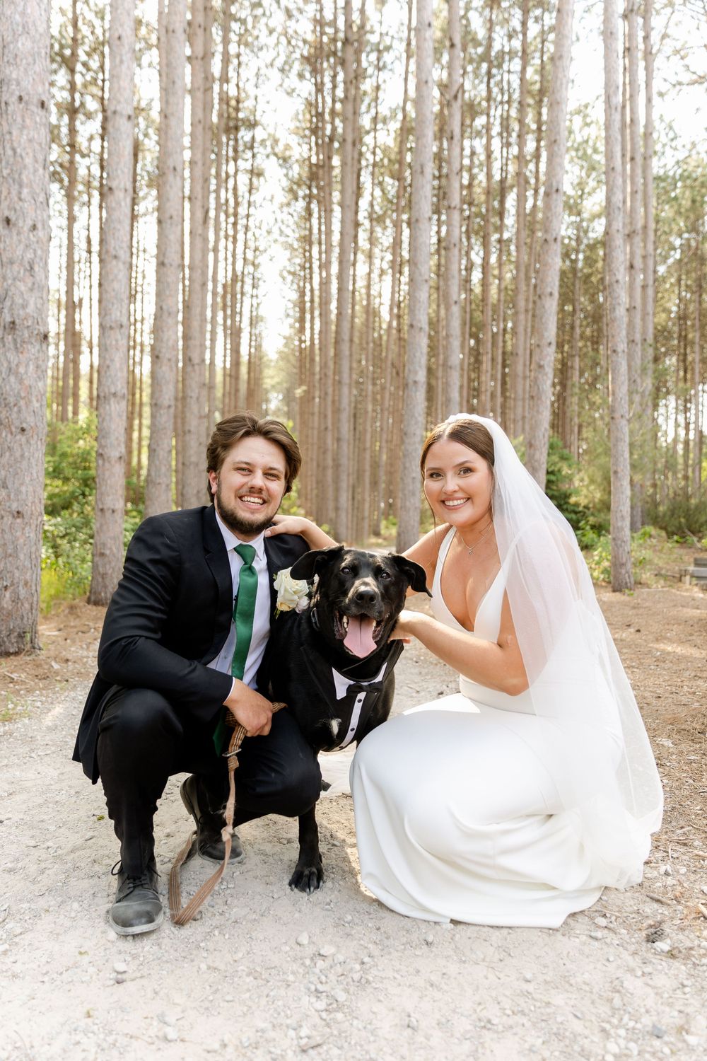 Happy couple in wedding attire posing with their black dog in a pine forest.