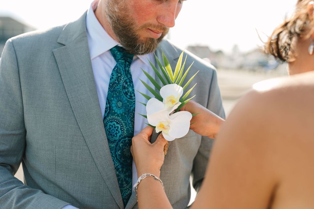 Close-up of boutonniere being pinned on gray suit jacket at wedding ceremony.