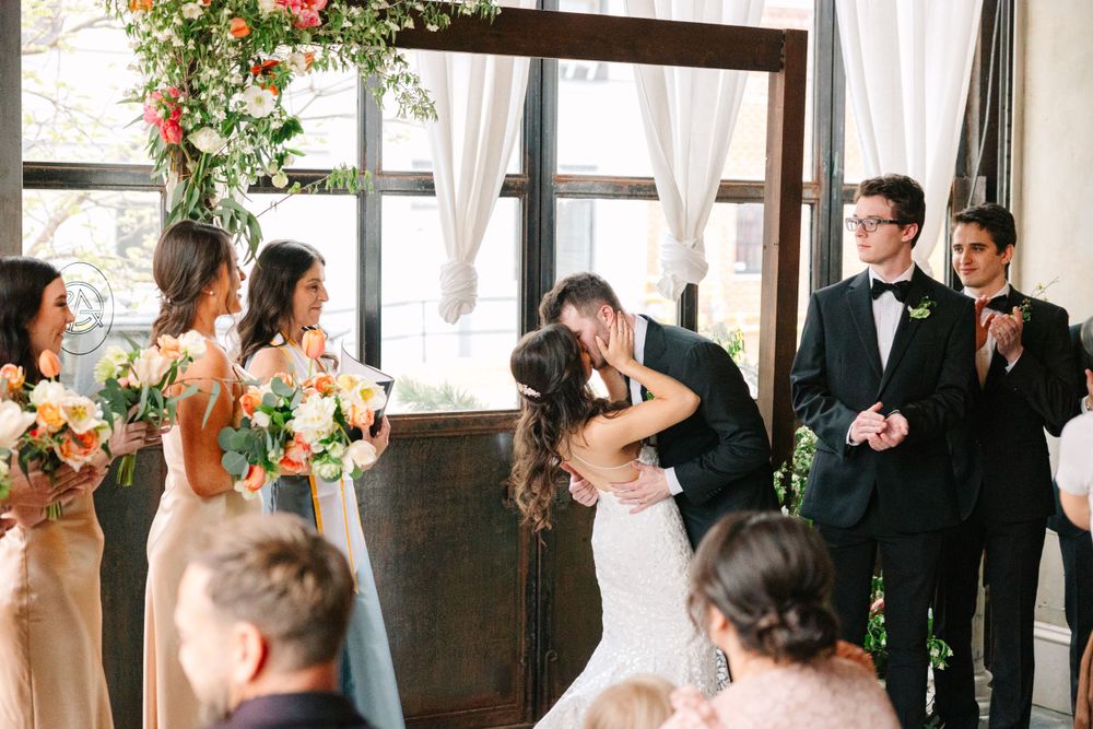 Wedding ceremony moment with couple kissing while wedding party looks on in indoor venue.