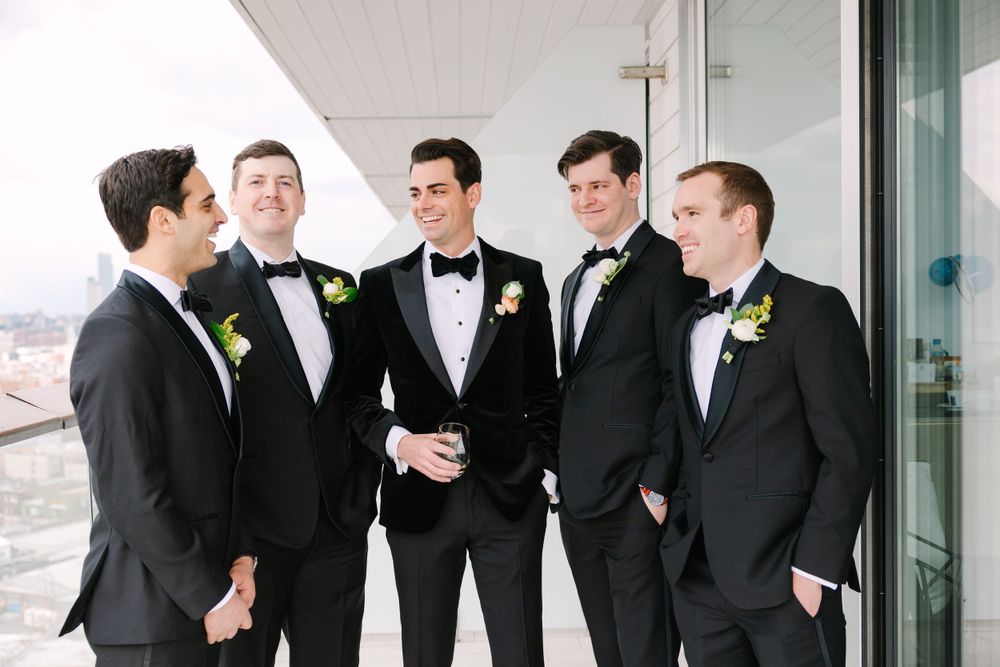 Groomsmen in black tuxedos sharing a candid moment on a balcony.
