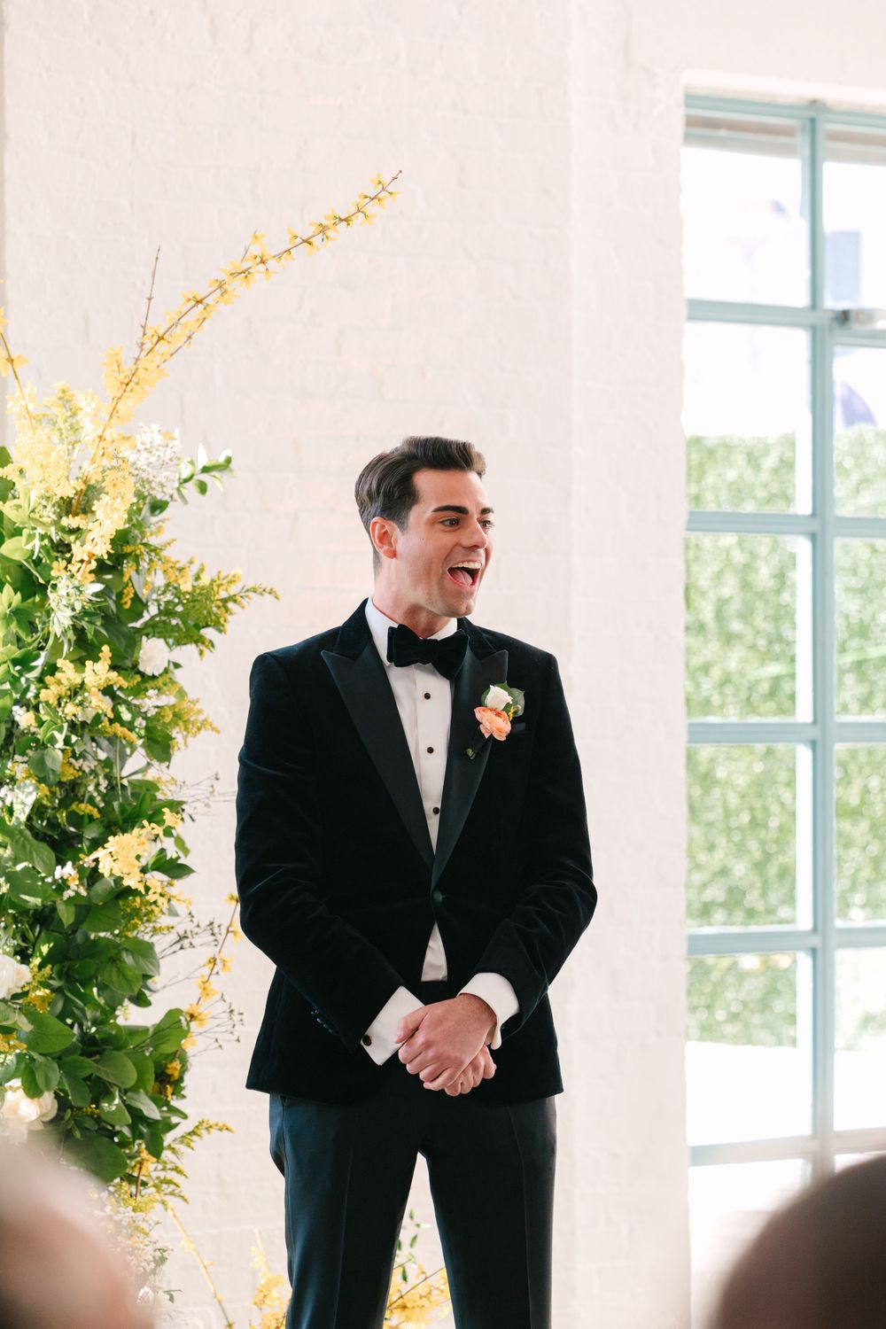 Groom in classic tuxedo smiling next to elegant floral arrangement.
