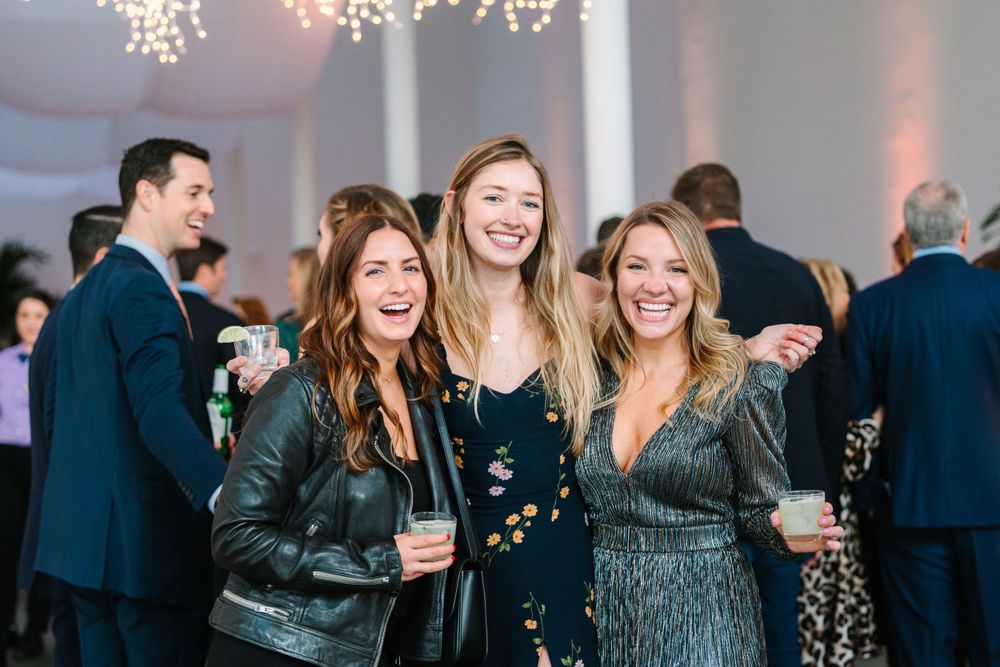 Three friends laughing together at an evening reception with twinkling lights.