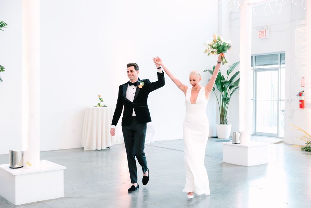 Couple in wedding attire joyfully entering their reception with raised hands.