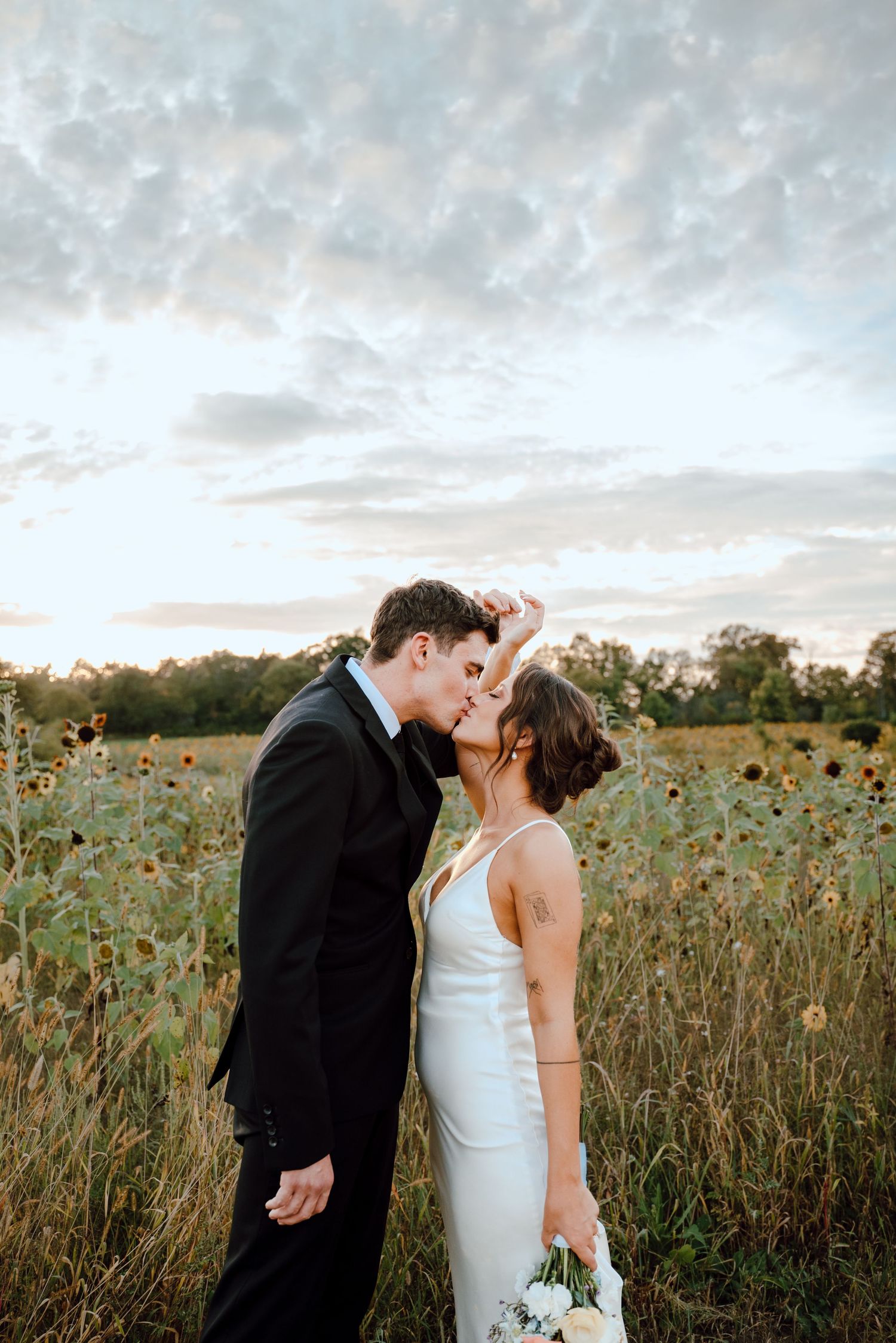 Romantic wedding couple embraces in a golden sunflower field at sunset with cloudy sky overhead.