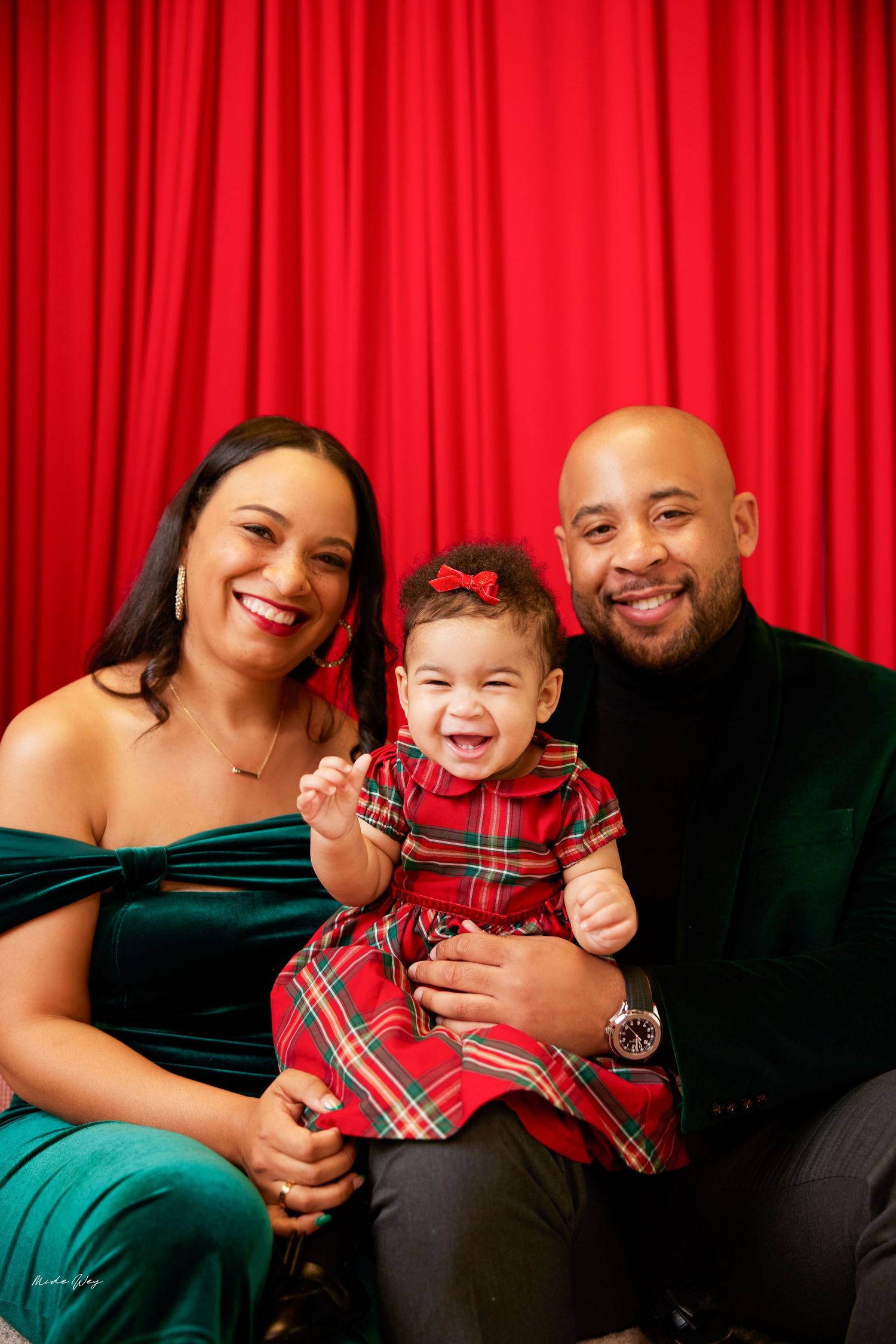 A happy family wearing red plaid and green outfits poses together for holiday photos against a red curtain backdrop.