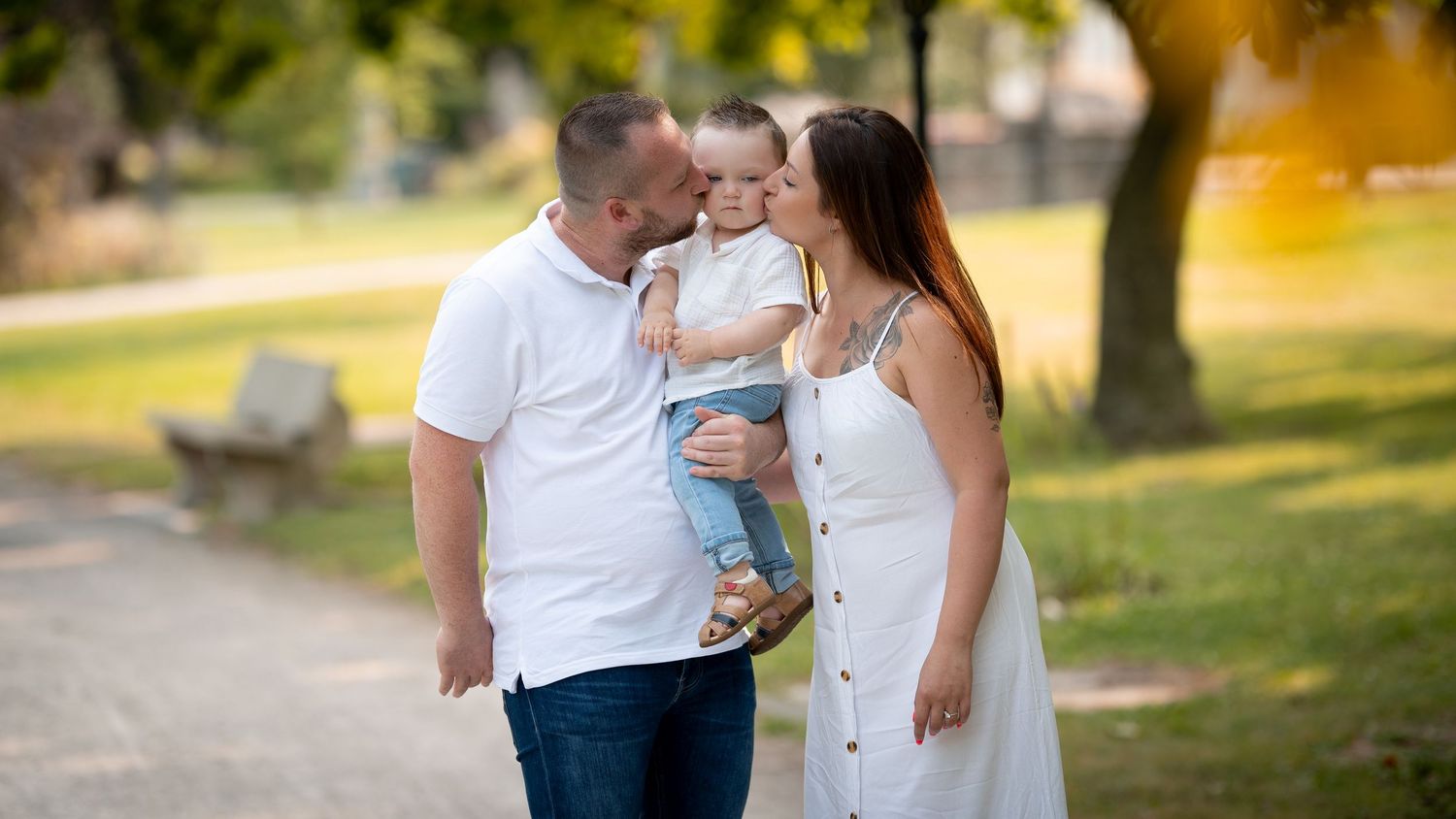 Loving parents sharing a tender moment as father holds baby close for a kiss in a sunlit park.