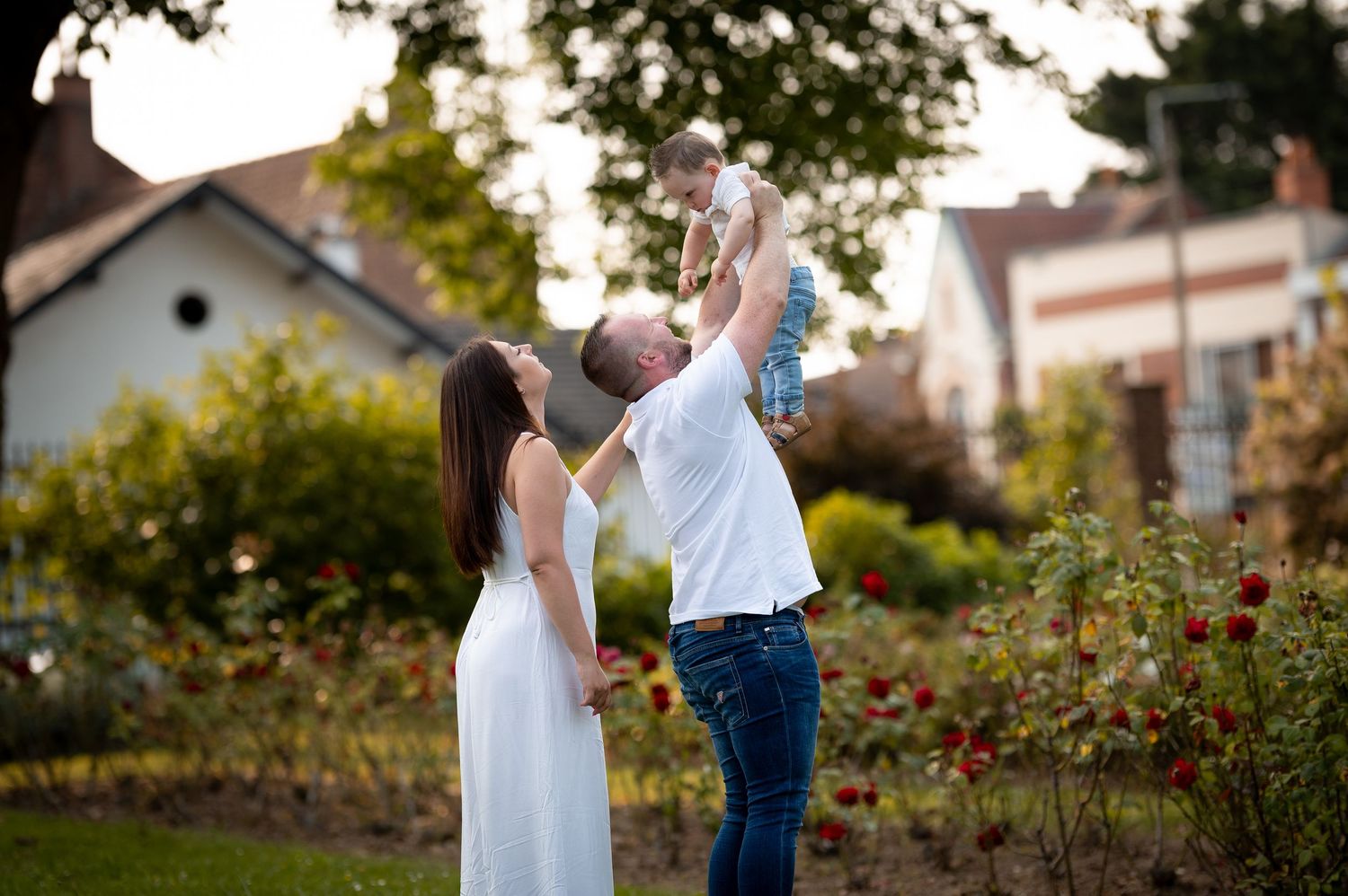Joyful father lifting baby high in the air while mother watches lovingly in a colorful garden setting.