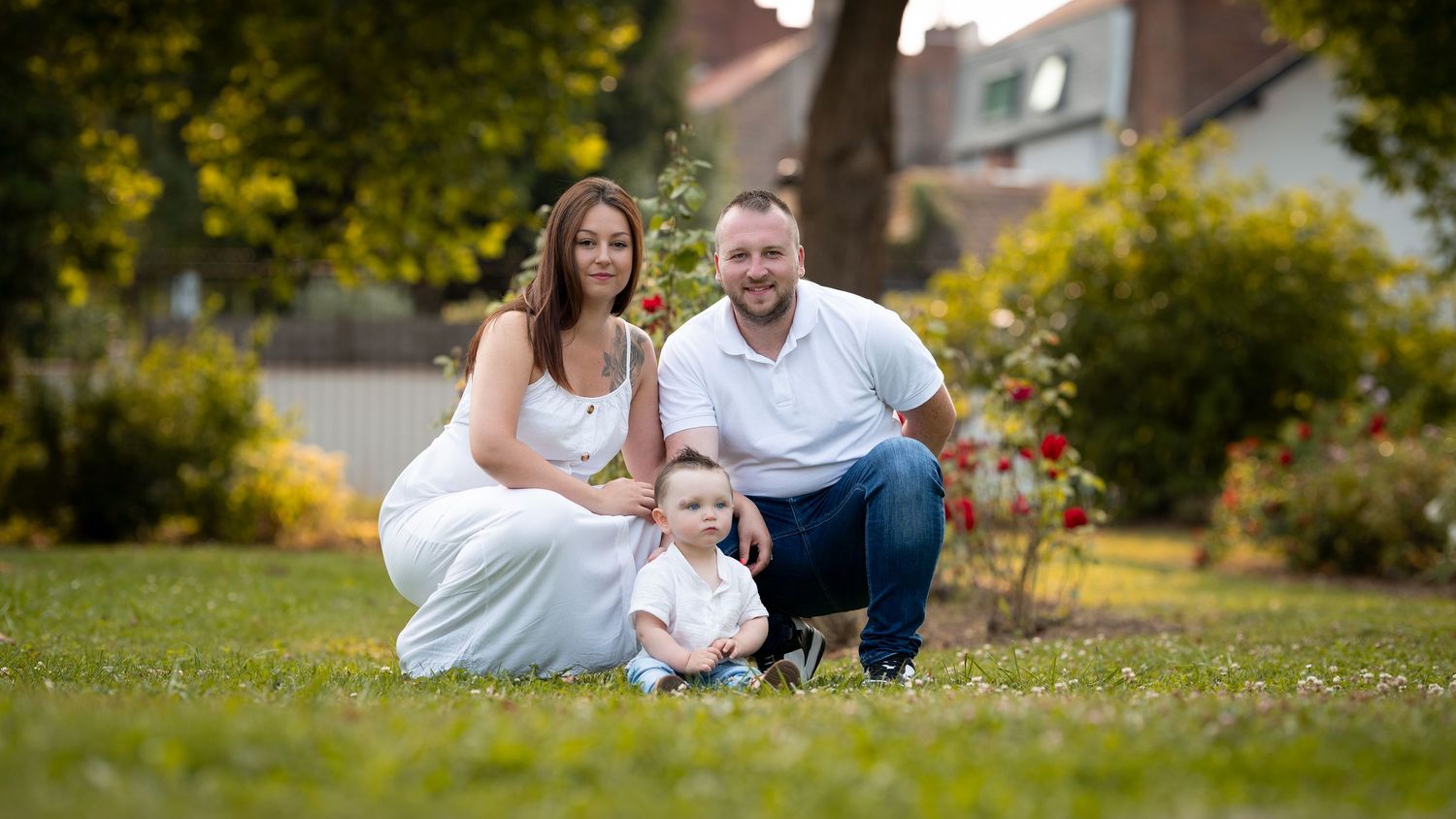 Happy family of three posing together on green grass with rose garden and house in the background.