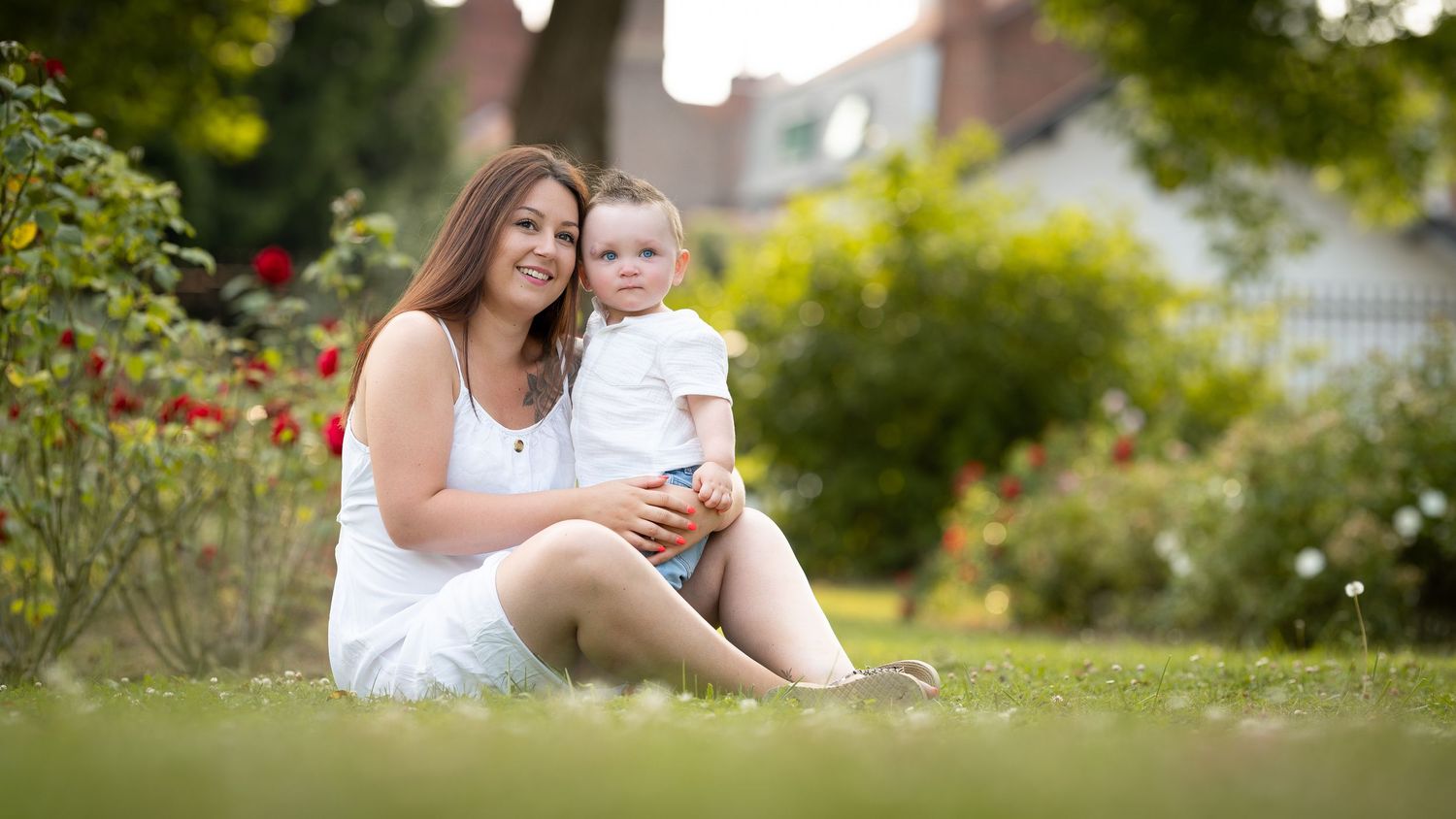 Smiling mother sitting on grass holding her young toddler in a beautiful garden with red roses.