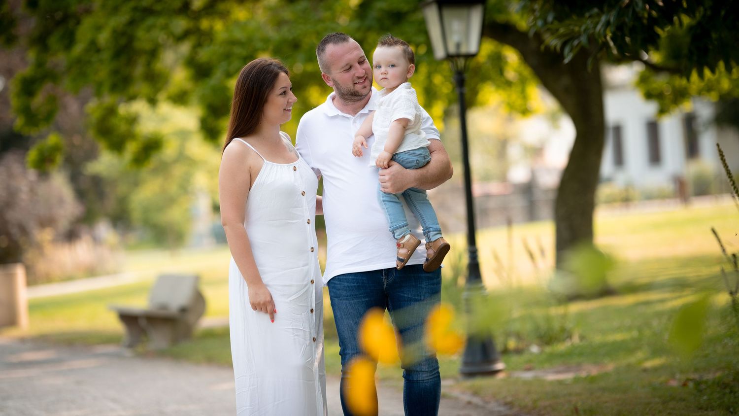 Happy young family with baby strolling along a sunny park pathway surrounded by lush green trees.