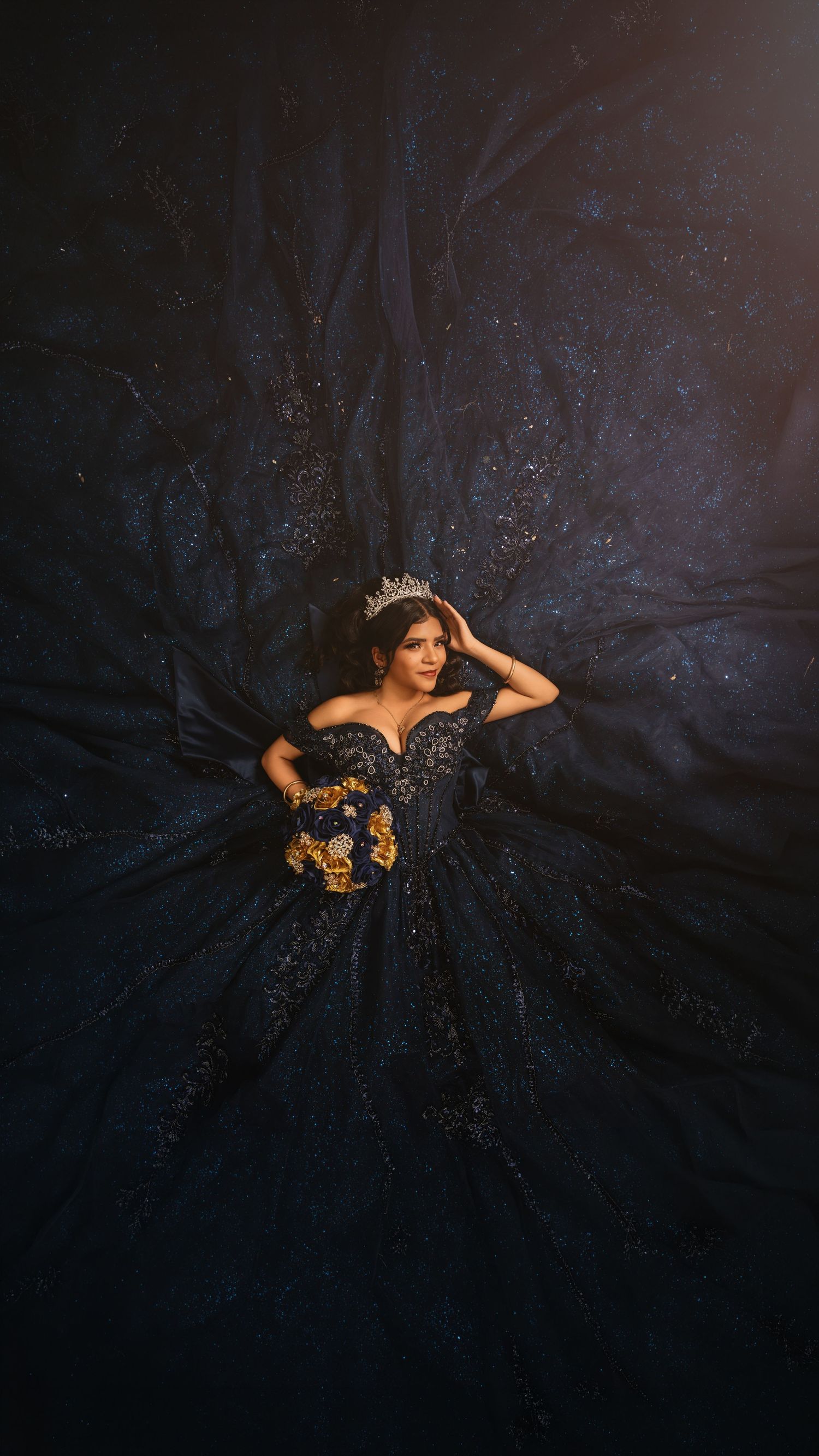 Young woman in navy blue ballgown with gold floral details and tiara poses elegantly with dress spread wide on dark background.