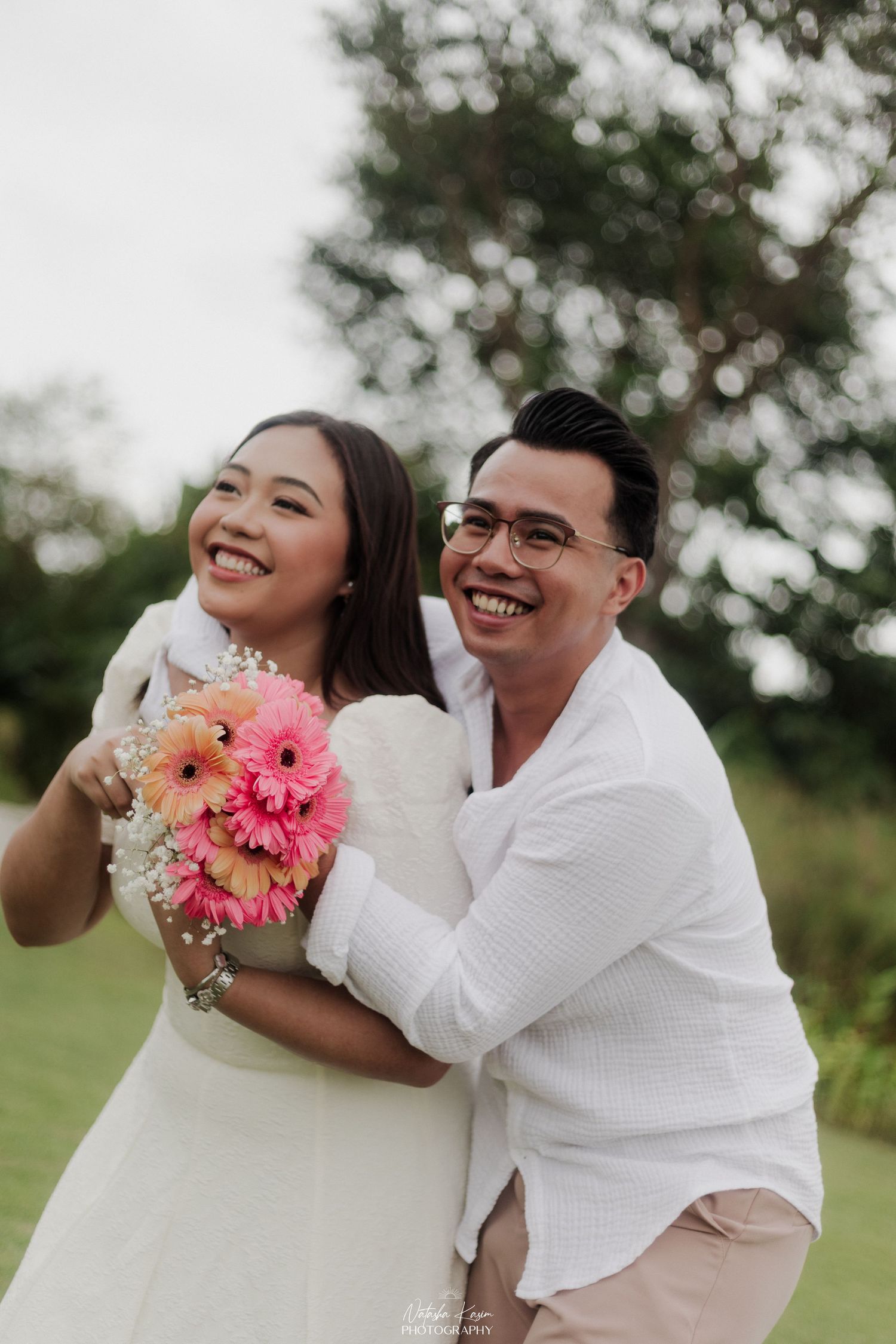 Happy couple in white outfits embracing outdoors, woman holding pink gerbera daisy bouquet, smiling joyfully.