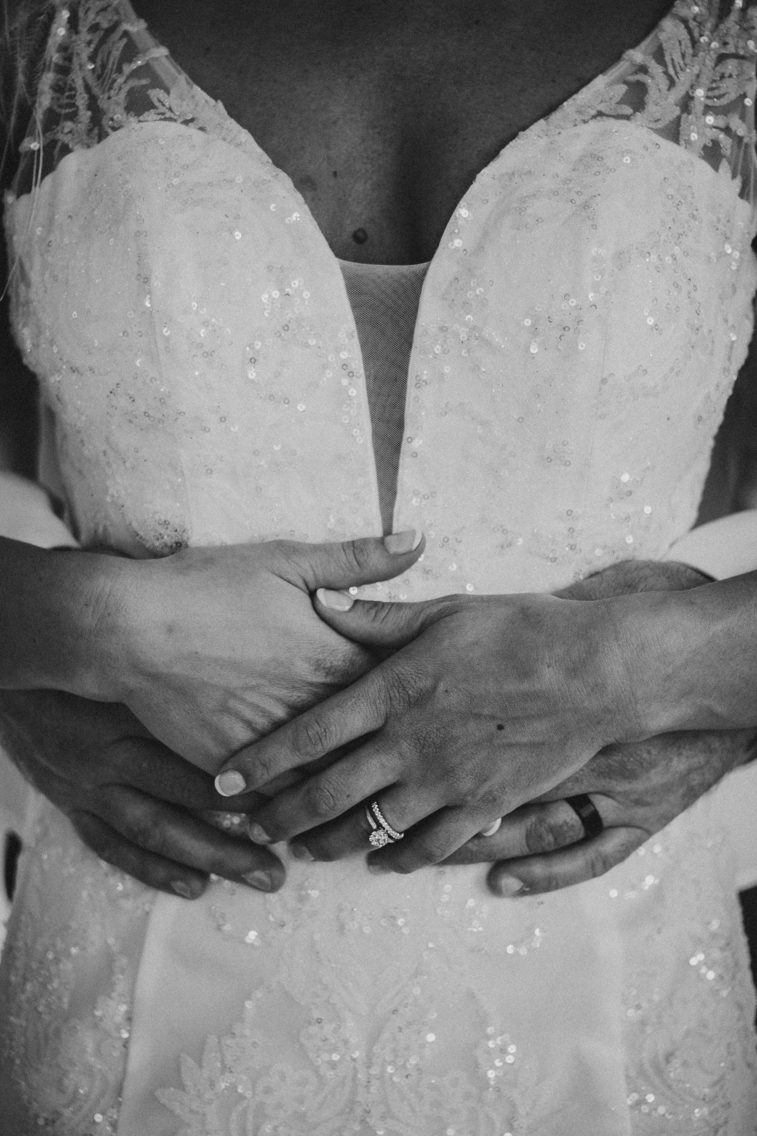 Close-up black and white photo of hands wearing wedding rings against a detailed white lace wedding dress.
