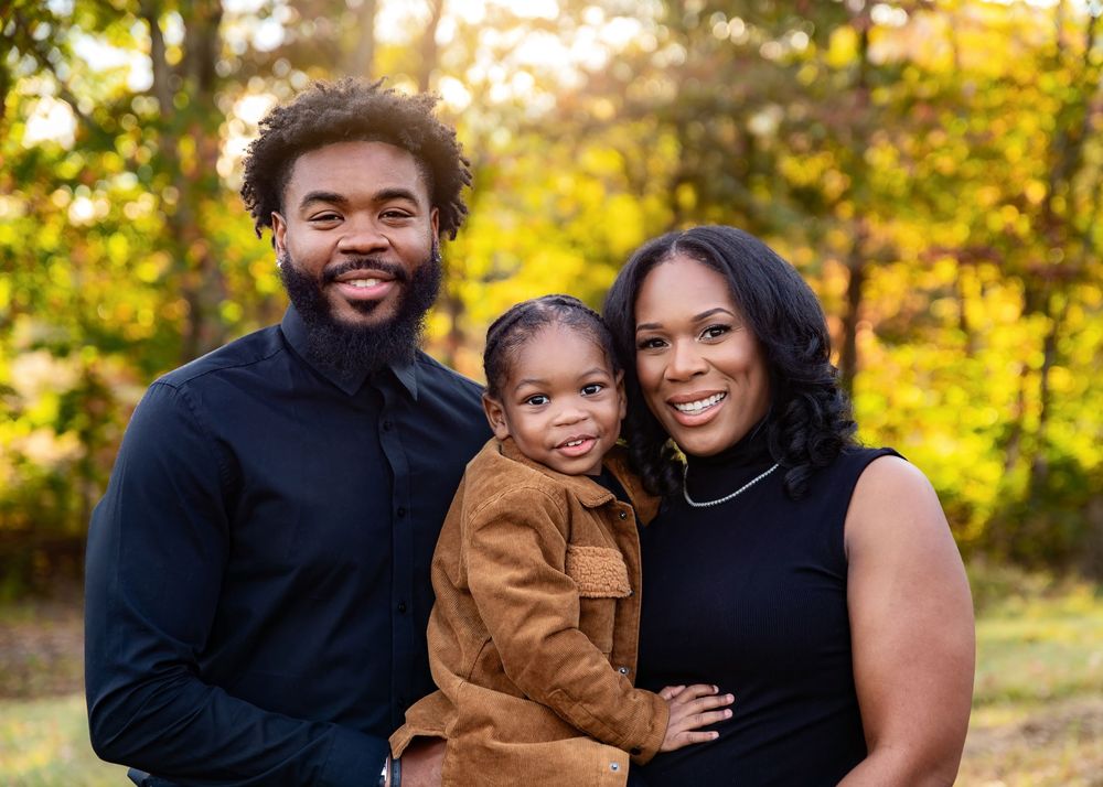 A happy Black family of three poses outdoors in fall, with parents smiling and holding their young toddler in a brown jacket.
