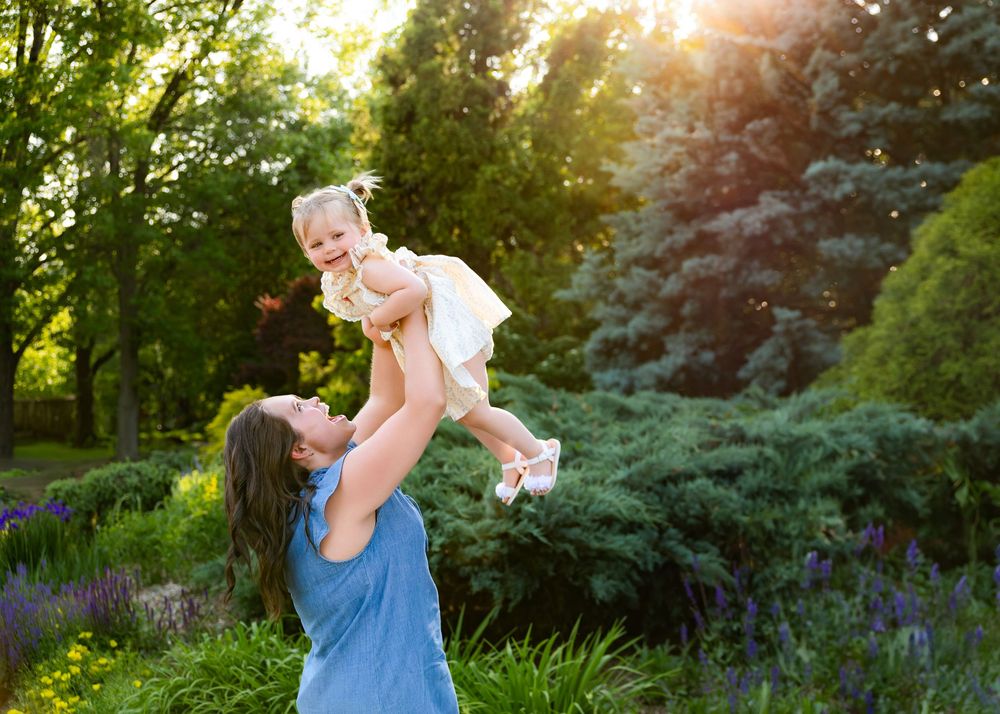 Sun glows through evergreen trees as two people dance and interact playfully in a lush summer garden setting.