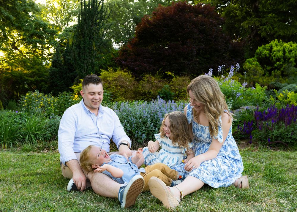 Family of four sitting on green grass laughing together in a lush garden with colorful flowers.
