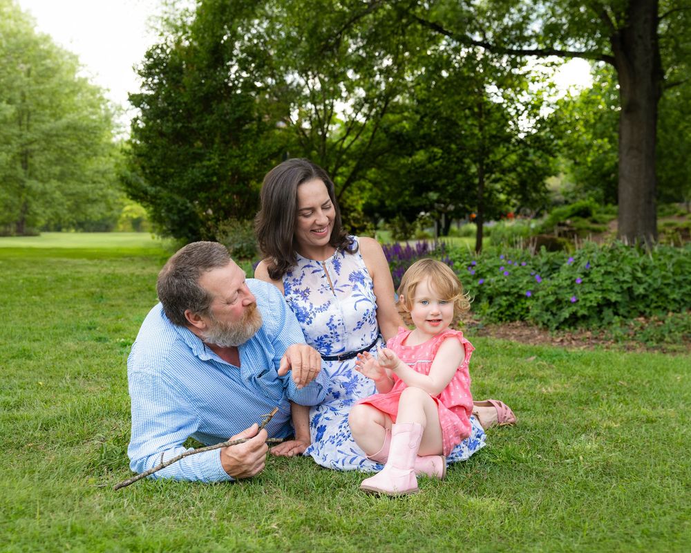 Family of three sitting on green grass in a park, parents smiling at toddler girl in pink dress on sunny day.