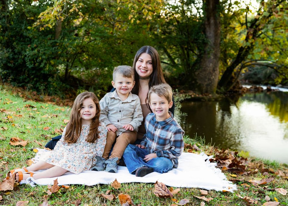 A smiling woman sits outdoors on a blanket with three young children surrounded by autumn leaves near a creek.