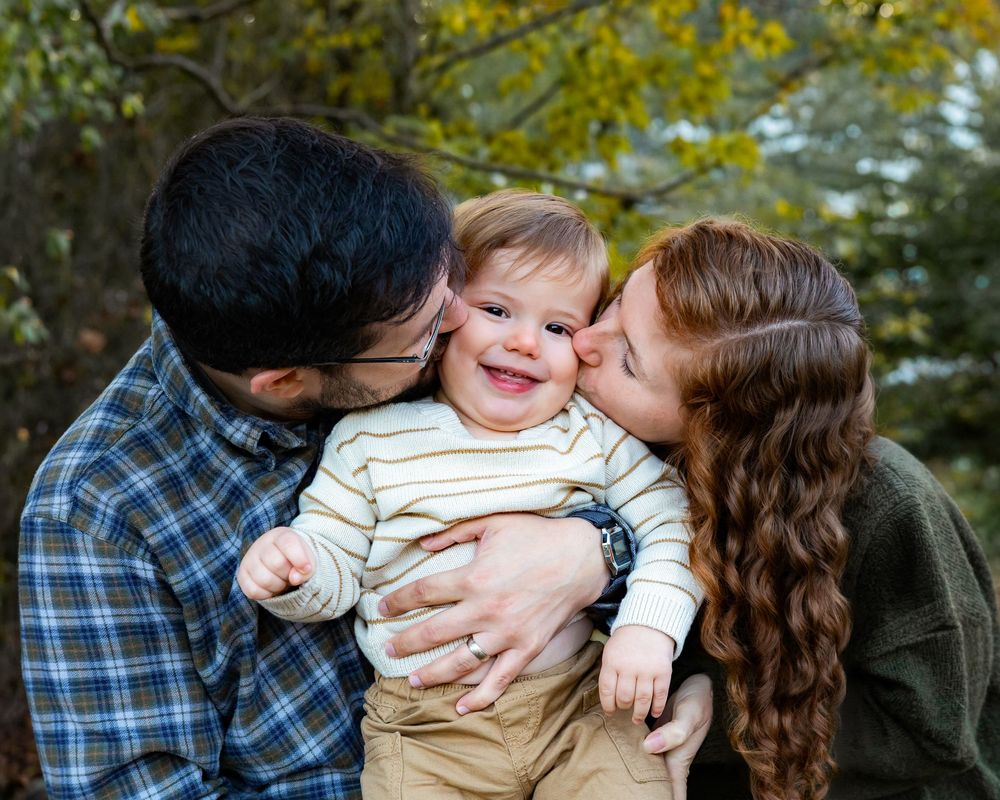 Happy toddler boy smiling while parents kiss his cheeks outdoors in autumn setting.