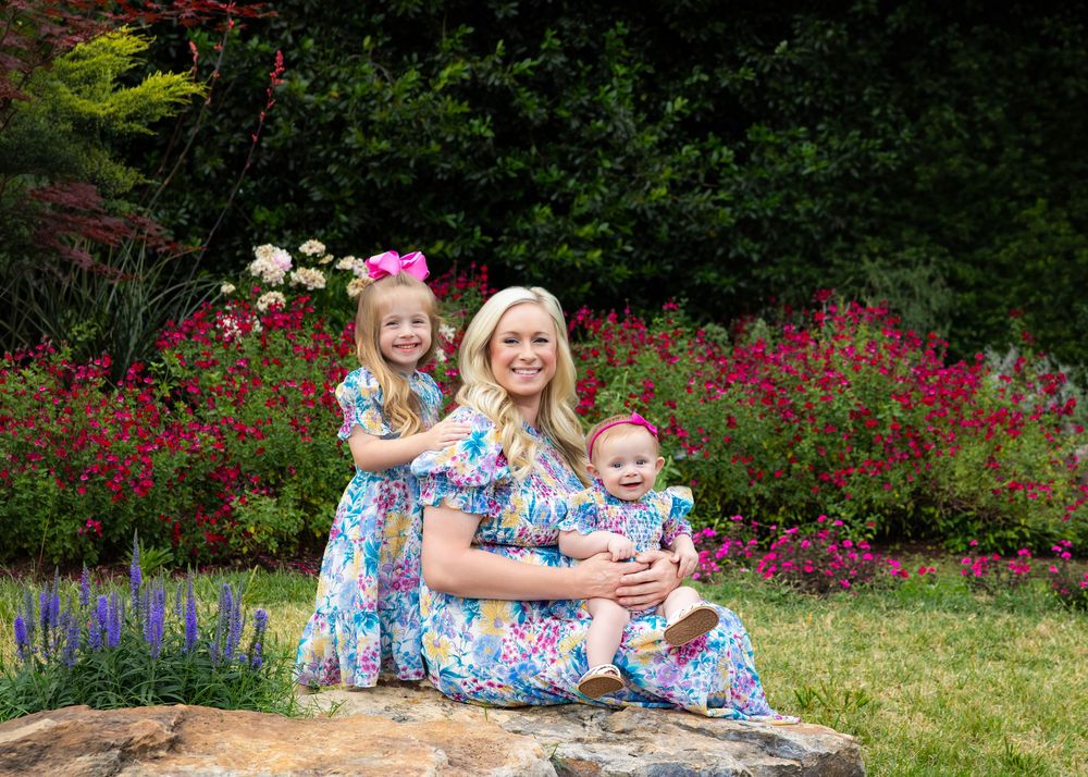 Mother and two daughters in matching floral dresses posing outdoors with colorful garden flowers in background.