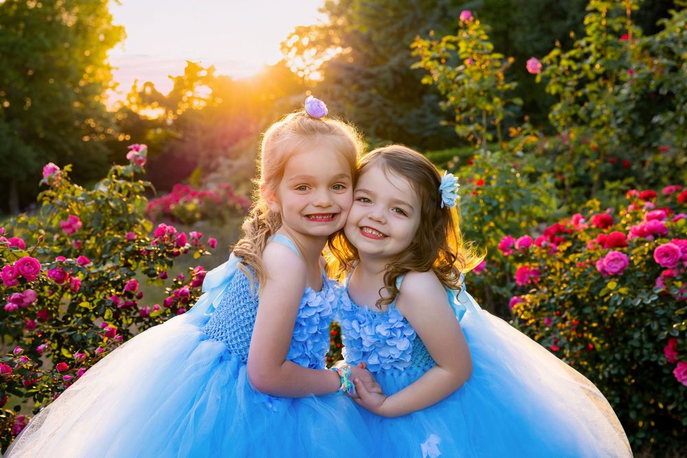 Two young girls in blue tutu dresses smile together among blooming roses in golden sunlight.