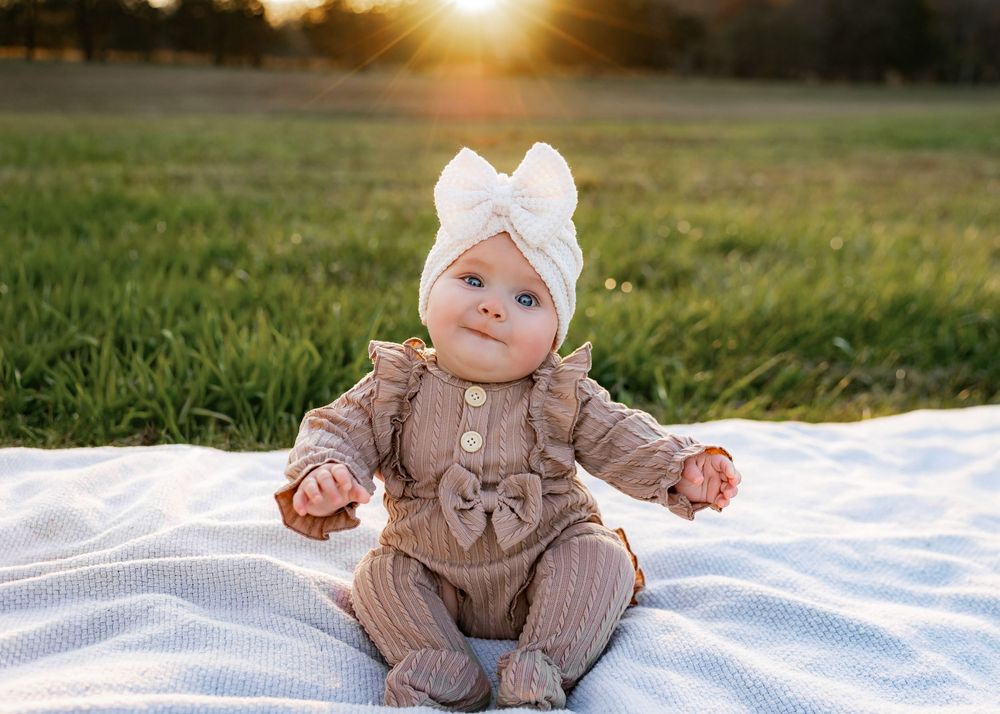 Baby wearing brown knit sweater and white hat sits on white blanket in grassy field at sunset in Knoxville, TN