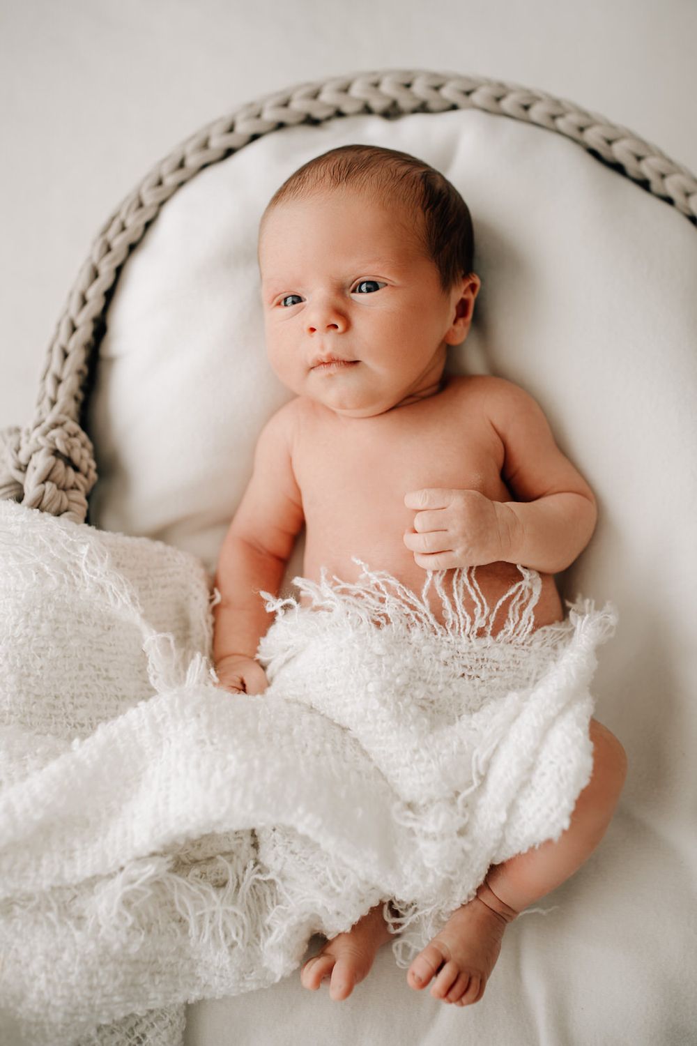 Baby nestled on white blanket with fringe detail against light gray knit backdrop during newborn photoshoot.Newborn Fotografie Frankfurt