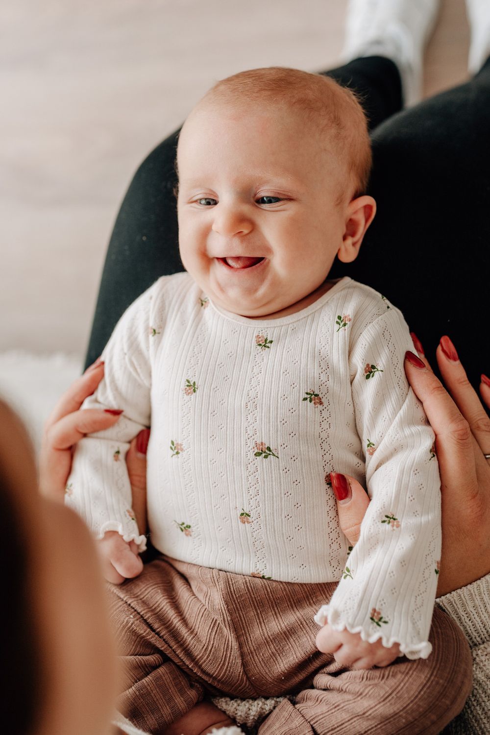 Baby wearing cream colored long sleeve sweater with small floral pattern and brown pants smiles joyfully at camera.Newborn Fotografie Frankf