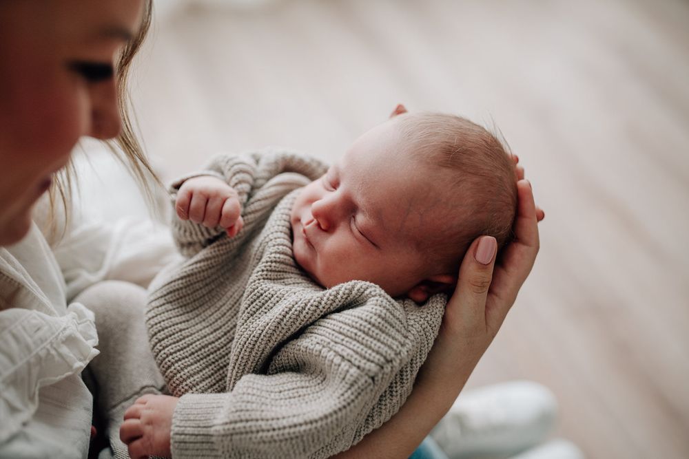 A sleeping newborn baby wearing a gray knit sweater is cradled gently in soft light.Newborn Fotografie Frankfurt