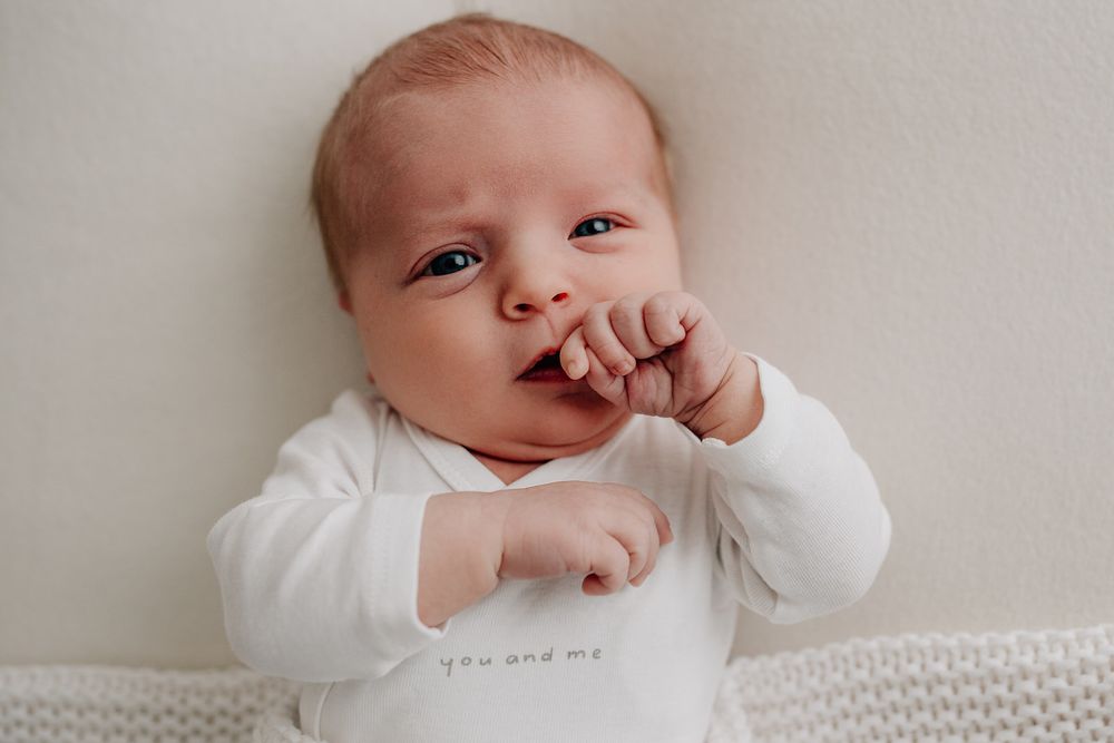 Baby wearing white onesie lying on a knitted blanket with hand near mouth.Newborn Fotografie Frankfurt