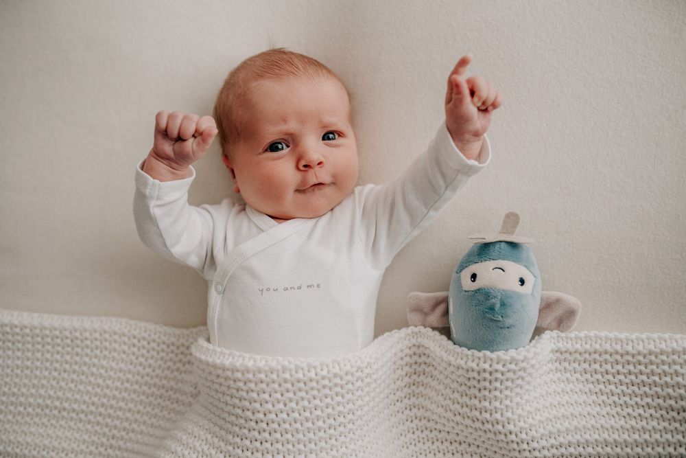 A baby with arms raised lies on white knit blanket next to a blue plush toy.Newborn Fotografie Frankfurt