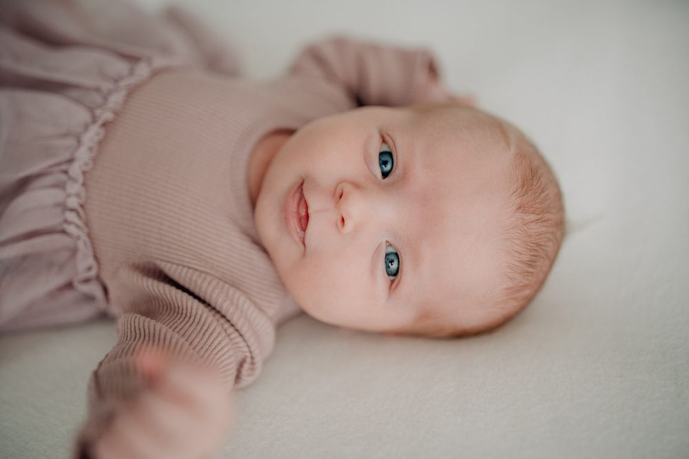 Baby with bright blue eyes wearing a mauve sweater smiles while lying on white bedding.Newborn Fotografie Frankfurt